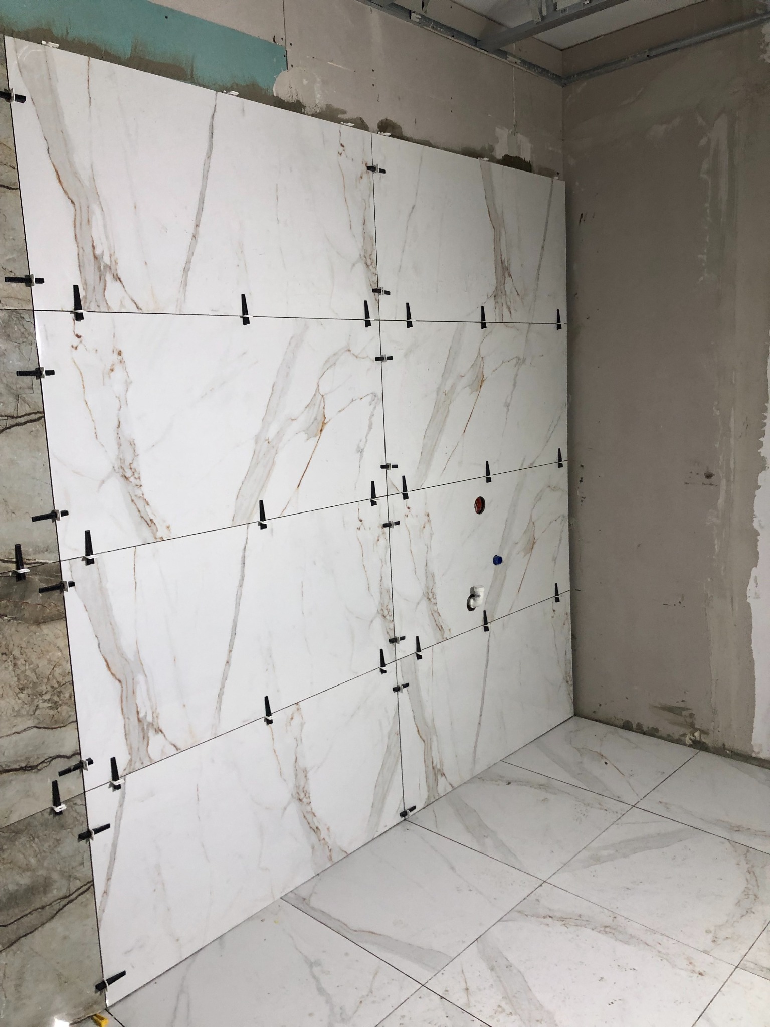 Układanie płytek imitujących marmur na ścianie i podłodze, z użyciem klipsów poziomujących, widoczne otwory na instalacje sanitarne.