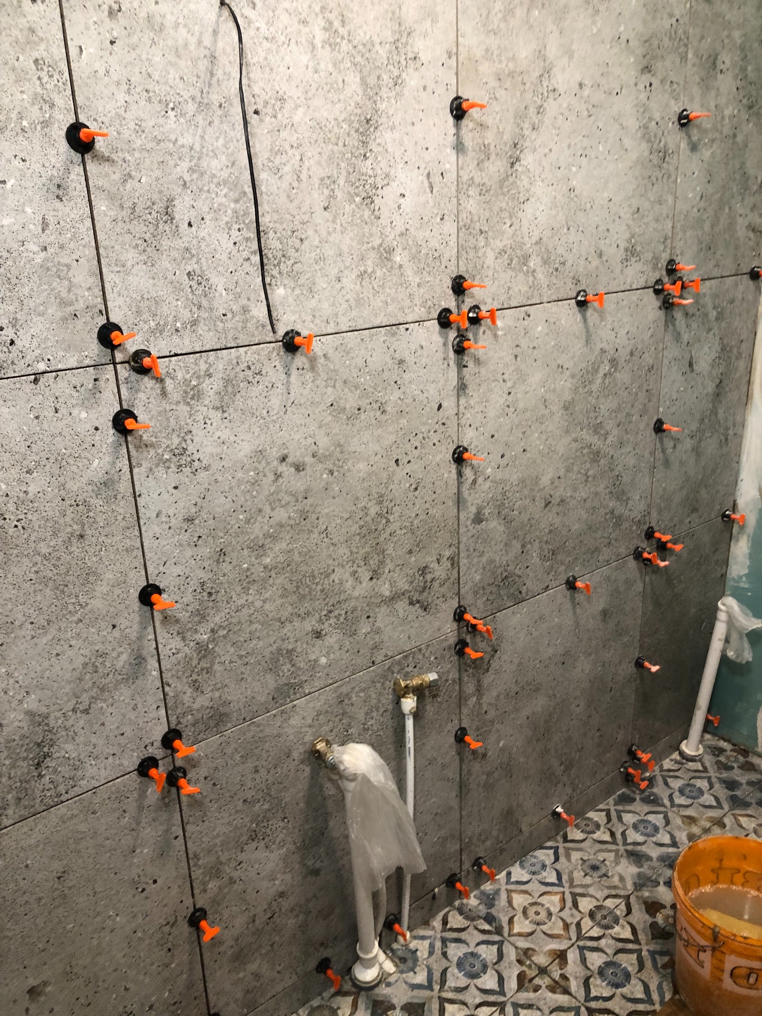 Szare płytki ścienne w trakcie montażu z użyciem klipsów poziomujących i krzyżyków dystansowych, widoczne rury instalacyjne i fragment wzorzystej podłogi.