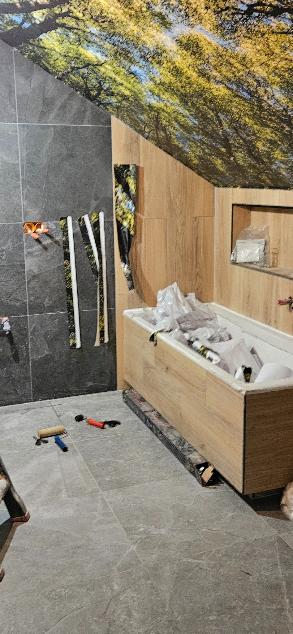 Niedokończona łazienka z szarymi płytkami, drewnopodobną zabudową wanny i fototapetą z motywem drzew, widoczne narzędzia glazurnicze na podłodze i rury wystające ze ściany.