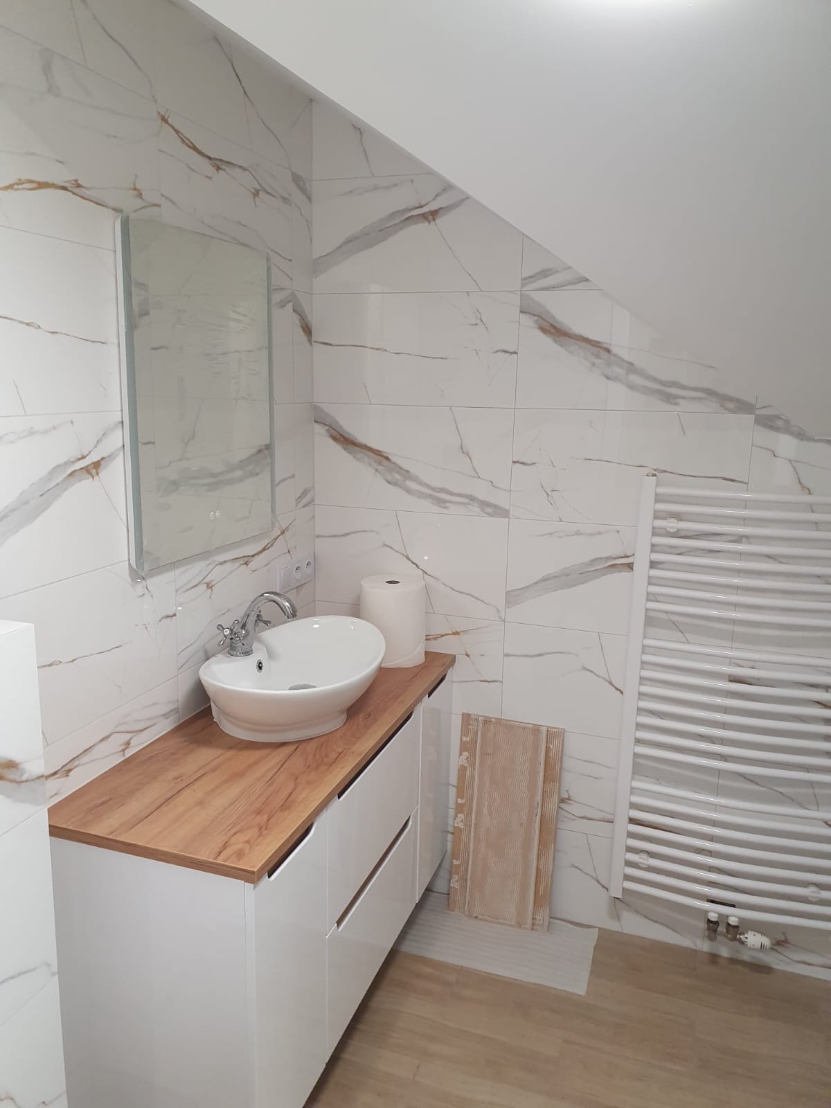 Wykończona łazienka na poddaszu: białe płytki imitujące marmur, drewnopodobny blat z owalną umywalką i grzejnik drabinkowy. Ukośne ściany.