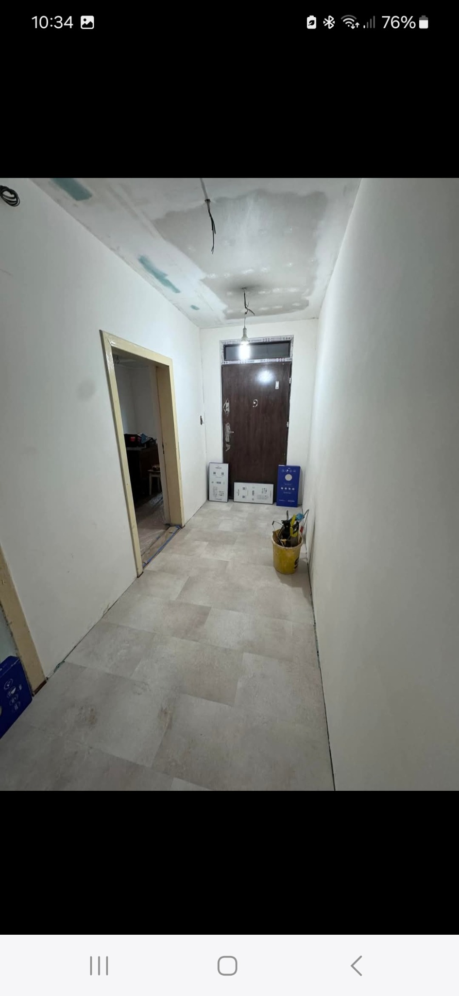 Korytarz w trakcie prac wykończeniowych: ściany po gładzi, ułożone panele podłogowe, sufit z widocznymi poprawkami, narzędzia w wiadrze. Widok na drzwi wejściowe.