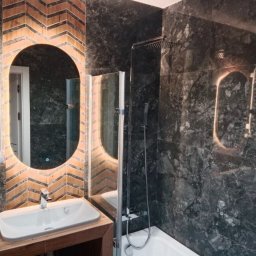 REMONTY I WYKOŃCZENIA NA TWOJĄ KIESZEŃ Małopolska i Śląsk - Nowoczesna łazienka z czarnymi marmurowymi płytkami, oświetlonym owalnym lustrem nad białą umywalką na drewnianym blacie, oraz grzejnikiem drabinkowym po lewej stronie.