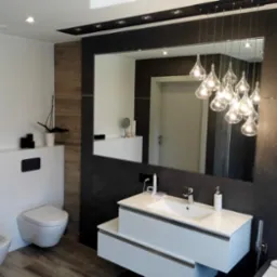 Nowoczesna łazienka z czarno-białą kolorystyką, podwieszaną toaletą i bidetem, dużym lustrem z oświetleniem w kształcie kropli oraz drewnopodobną podłogą.