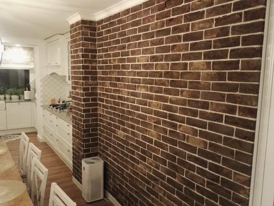 Ściana wykończona cegłą klinkierową w odcieniach brązu z jasną fugą, widok na białą kuchnię w tle, z widocznym oczyszczaczem powietrza.