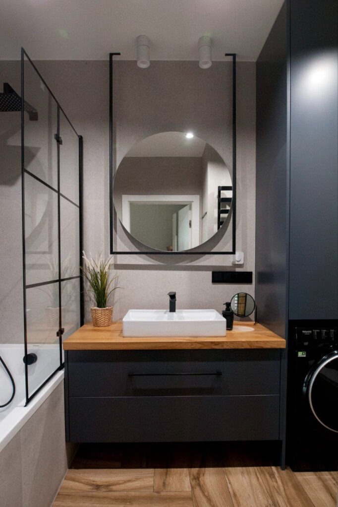 Nowoczesna łazienka z umywalką nablatową na drewnianym blacie, okrągłym lustrem w czarnej ramie i kabiną prysznicową ze szprosami.
