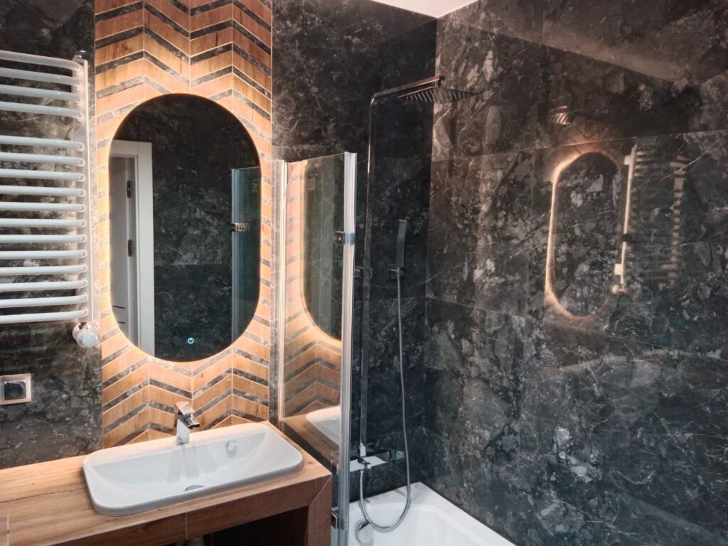 Nowoczesna łazienka z czarnymi marmurowymi płytkami, oświetlonym owalnym lustrem nad białą umywalką na drewnianym blacie, oraz grzejnikiem drabinkowym po lewej stronie.