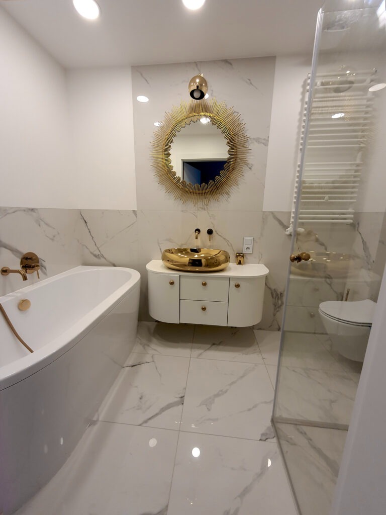 Biała łazienka z marmurowymi płytkami, złotą umywalką i lustrem w kształcie słońca, bateria wannowa w kolorze złota, kabina prysznicowa ze szklanymi drzwiami.
