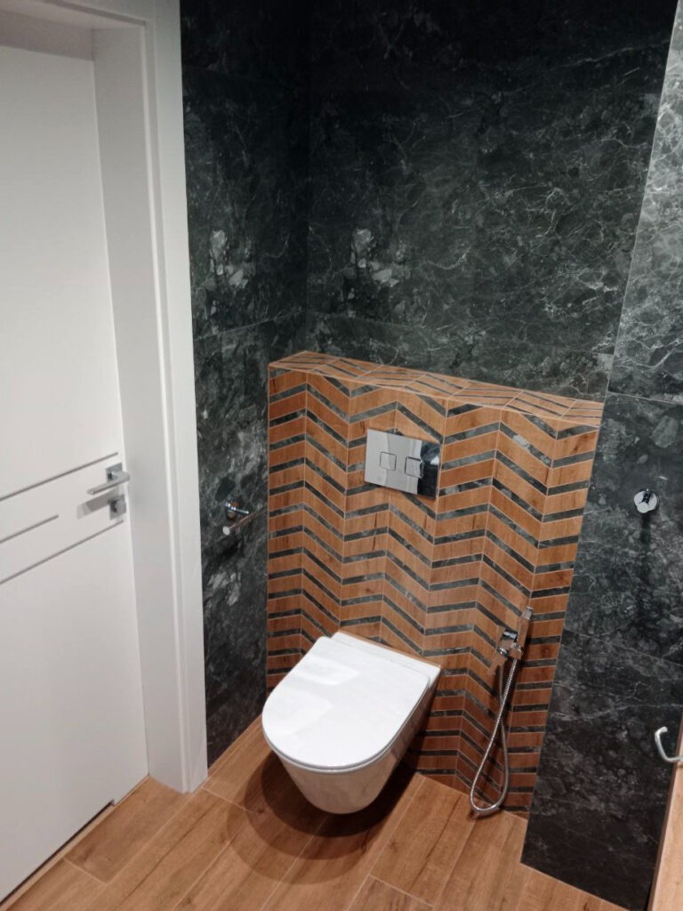 Nowoczesna toaleta podwieszana z deską wolnoopadającą, zamontowana na tle ściany wyłożonej płytkami w jodełkę w odcieniach brązu i szarości, sąsiadującej z ciemną, marmurkową ścianą i drewnianą...