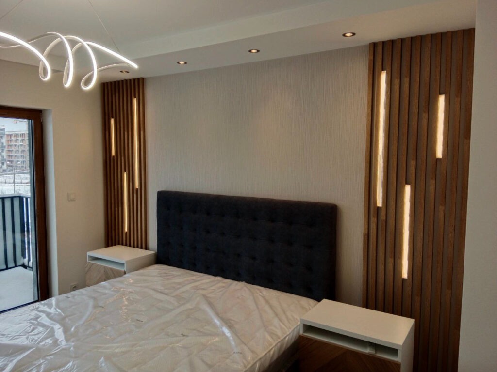 Nowoczesna sypialnia z drewnianymi panelami ściennymi z wbudowanym oświetleniem LED po bokach łóżka z ciemnym zagłówkiem, białe szafki nocne i futurystyczna lampa sufitowa.