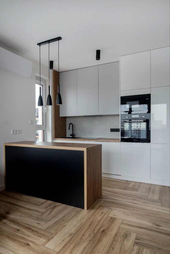 Nowoczesna kuchnia z białymi szafkami, drewnianym blatem, czarnym zlewem i wyspą z czarnym frontem i drewnianymi bokami, podłoga z paneli ułożonych w jodełkę.