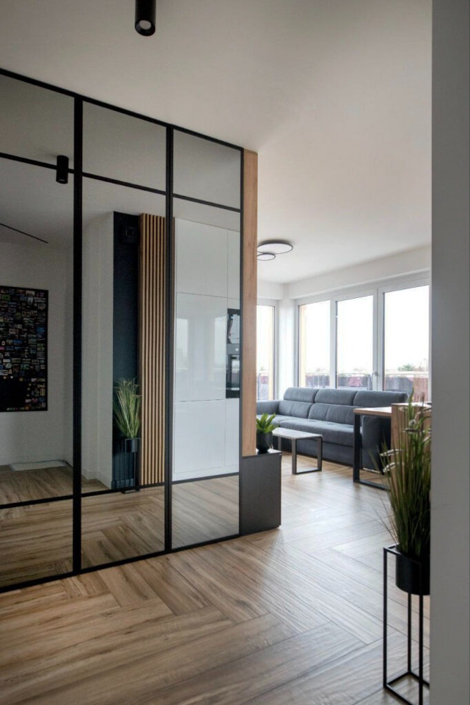 Nowoczesne wnętrze z drewnianą podłogą ułożoną w jodełkę, szklaną ścianą działową z czarnymi profilami, widok na kuchnię z białymi szafkami i salon z szarą sofą.