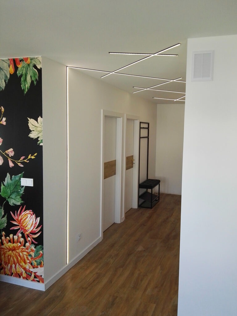 Korytarz z drewnopodobną podłogą, czarną fototapetą z motywem kwiatowym i geometrycznie ułożonymi liniami świetlnymi na suficie.