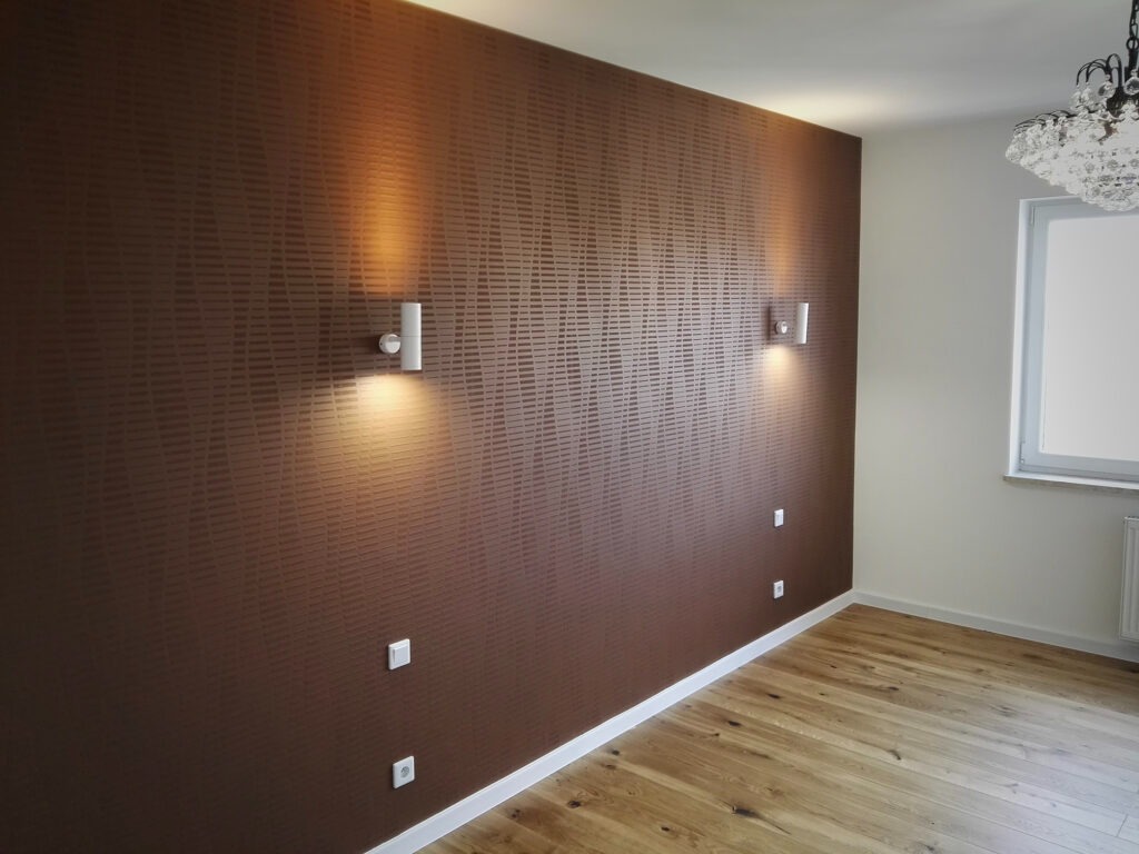 Brązowa ściana z wzorem, oświetlona dwoma kinkietami, parkiet z jasnego drewna, kryształowy żyrandol w rogu pokoju.