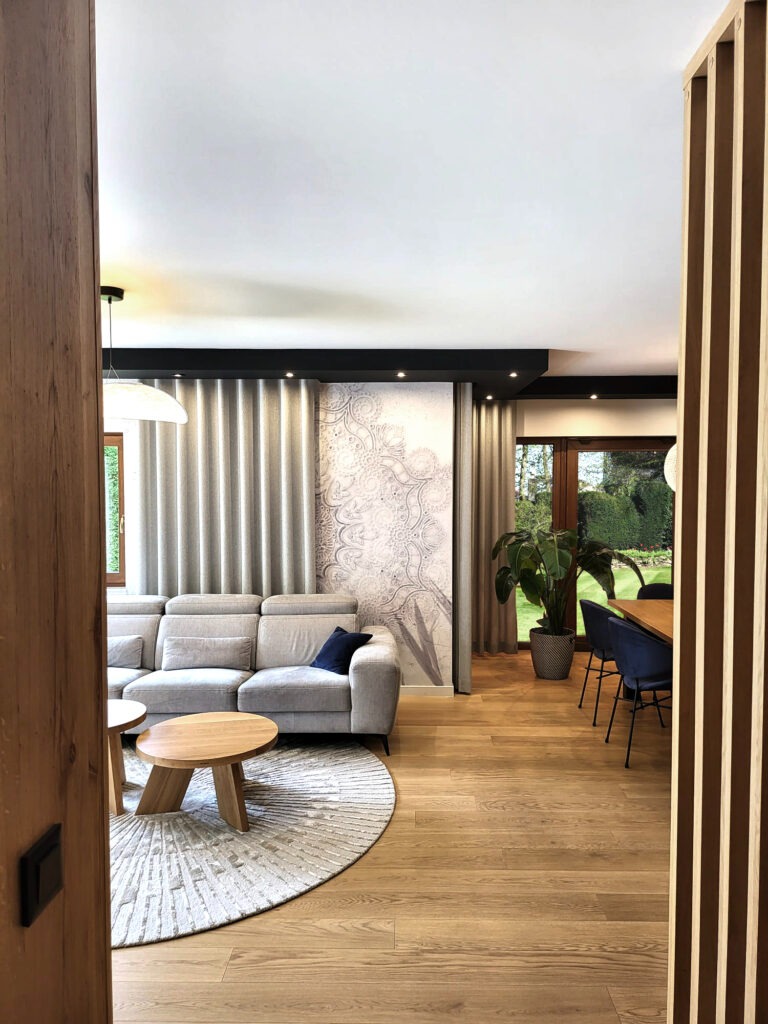 Jasny salon z drewnianą podłogą, szarą sofą z granatową poduszką, okrągłym dywanem, drewnianymi stolikami kawowymi i ścianą z dekoracyjnym wzorem.