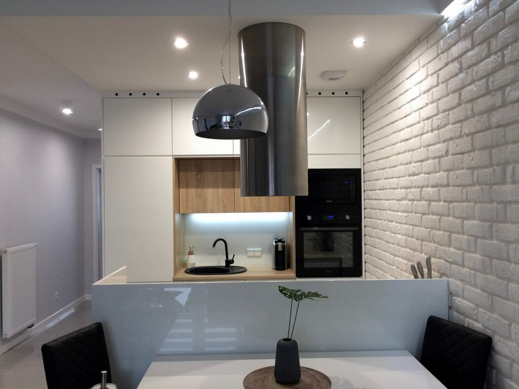 Nowoczesna kuchnia z białymi szafkami, drewnianymi akcentami, chromowanym okapem i lampą, białą ceglaną ścianą, oraz stołem z rośliną w wazonie.