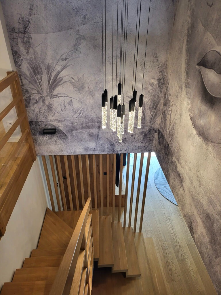 Drewniane schody z ażurową balustradą, oświetlone wiszącą lampą z kryształowymi elementami, na tle ściany z fototapetą przedstawiającą motywy roślinne i fragment twarzy.