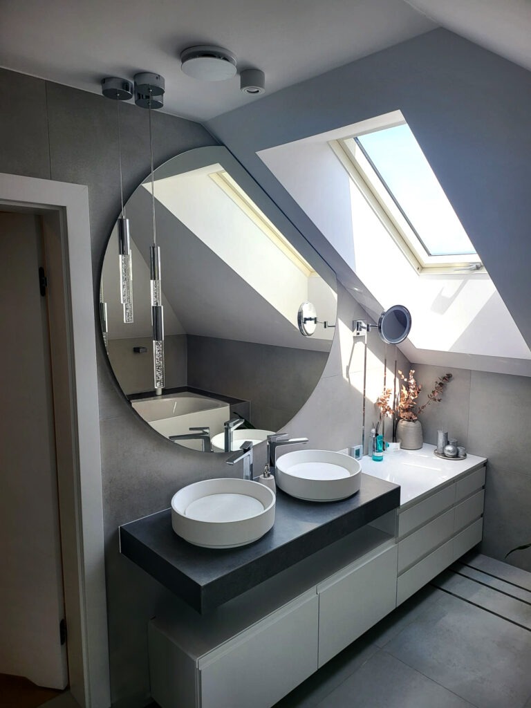 Nowoczesna łazienka na poddaszu z dwoma okrągłymi umywalkami nablatowymi, okrągłym lustrem i wiszącymi lampami kryształowymi, oświetlona naturalnym światłem wpadającym przez okno dachowe.