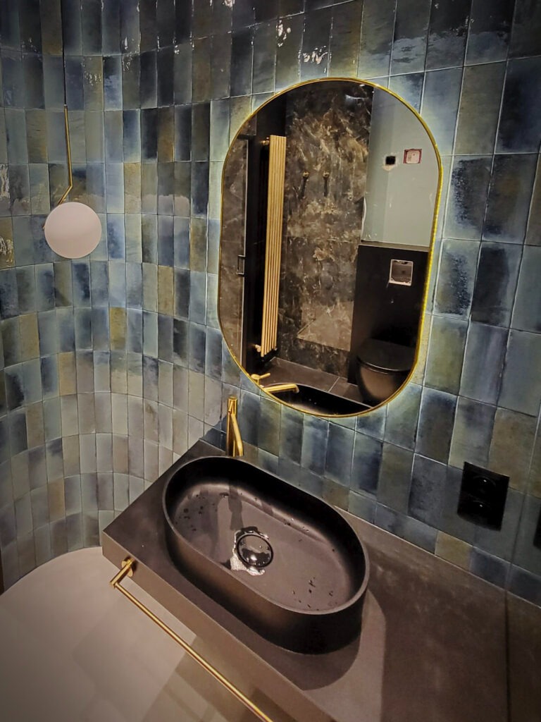 Nowoczesna łazienka z umywalką nablatową w kolorze czarnym, baterią w kolorze złotym i lustrem z podświetleniem LED w złotej ramie na tle ściany wyłożonej niebiesko-zielonymi płytkami.