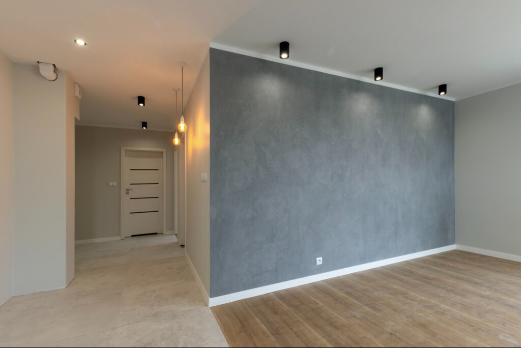 Nowoczesne wnętrze z szarą ścianą o fakturze betonu, drewnianą podłogą i minimalistycznym oświetleniem punktowym oraz wiszącymi lampami z żarówkami edisona.