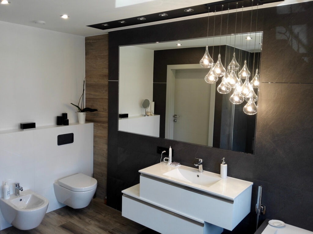 Nowoczesna łazienka z czarno-białą kolorystyką, podwieszaną toaletą i bidetem, dużym lustrem z oświetleniem w kształcie kropli oraz drewnopodobną podłogą.