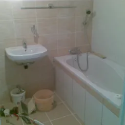 Łazienka w trakcie remontu: biała wanna obudowana płytkami, umywalka wisząca, narzędzia i wiadra na podłodze, widoczne niedokończone wykończenia przy framudze drzwi.