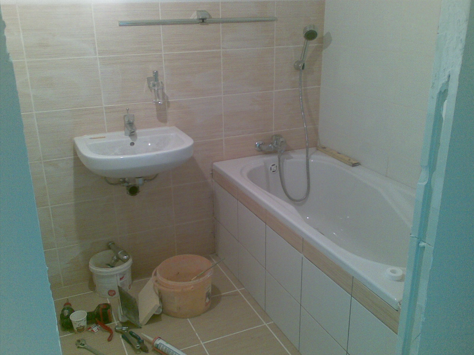 Łazienka w trakcie remontu: biała wanna obudowana płytkami, umywalka wisząca, narzędzia i wiadra na podłodze, widoczne niedokończone wykończenia przy framudze drzwi.