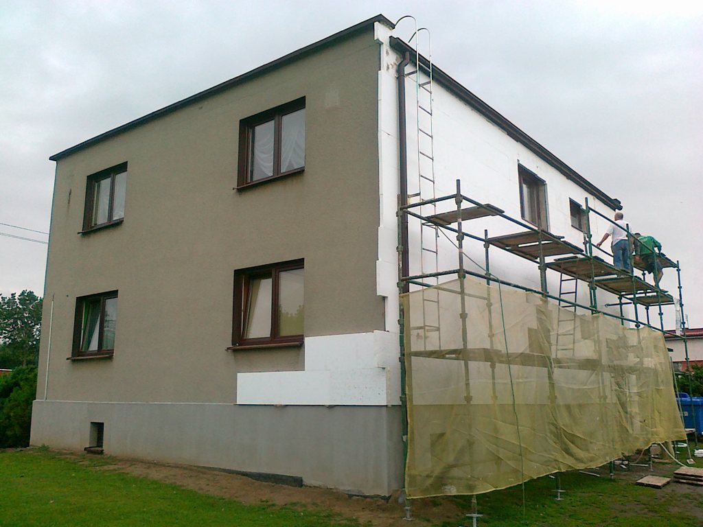 Dwukondygnacyjny dom w trakcie docieplania elewacji styropianem, widoczne rusztowanie, pracownicy w trakcie montażu, fragment ściany z szarym tynkiem.