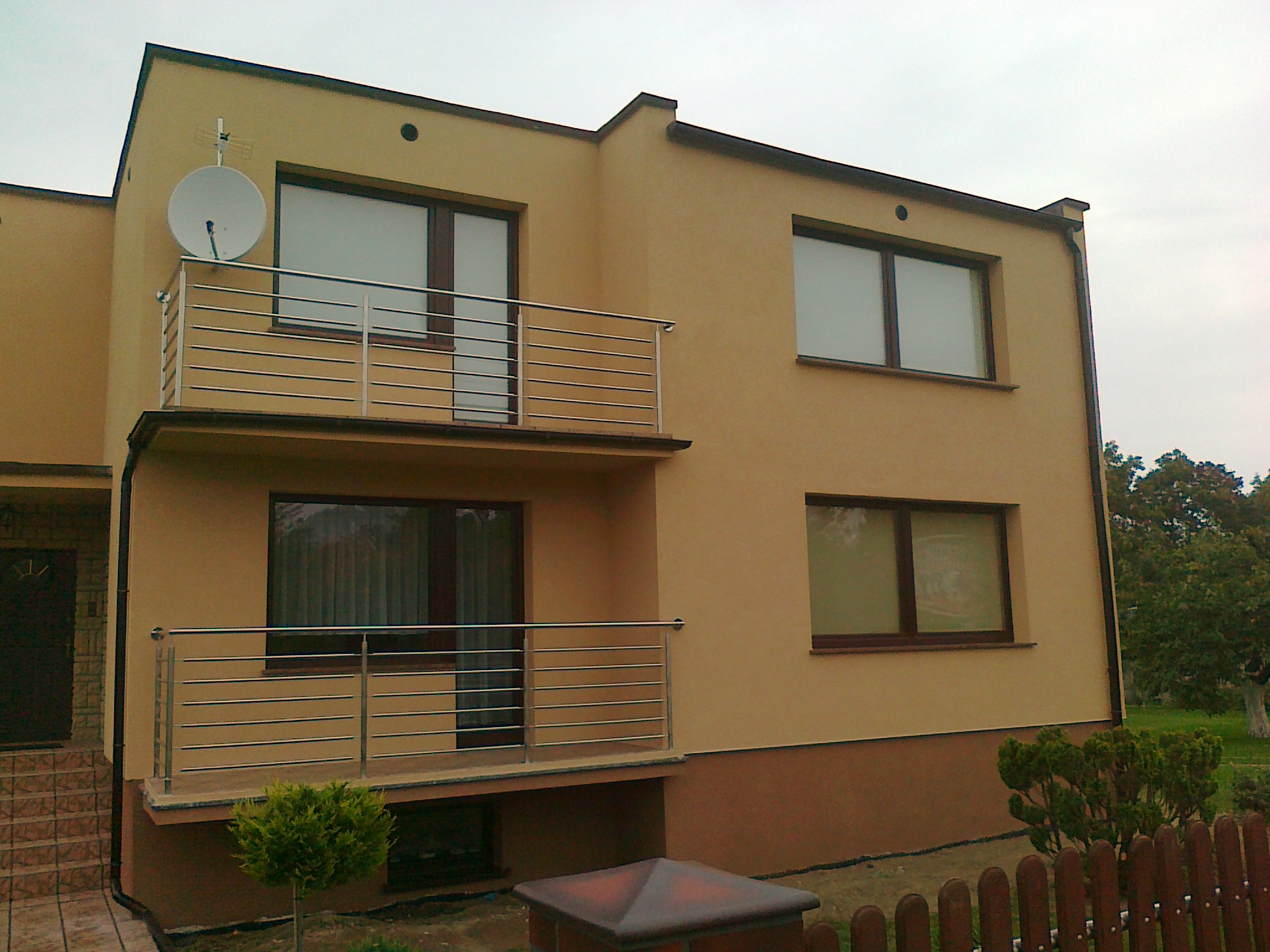 Dwupiętrowy dom z beżową elewacją, balkonami z metalowymi balustradami i anteną satelitarną na dachu, widok z poziomu gruntu.