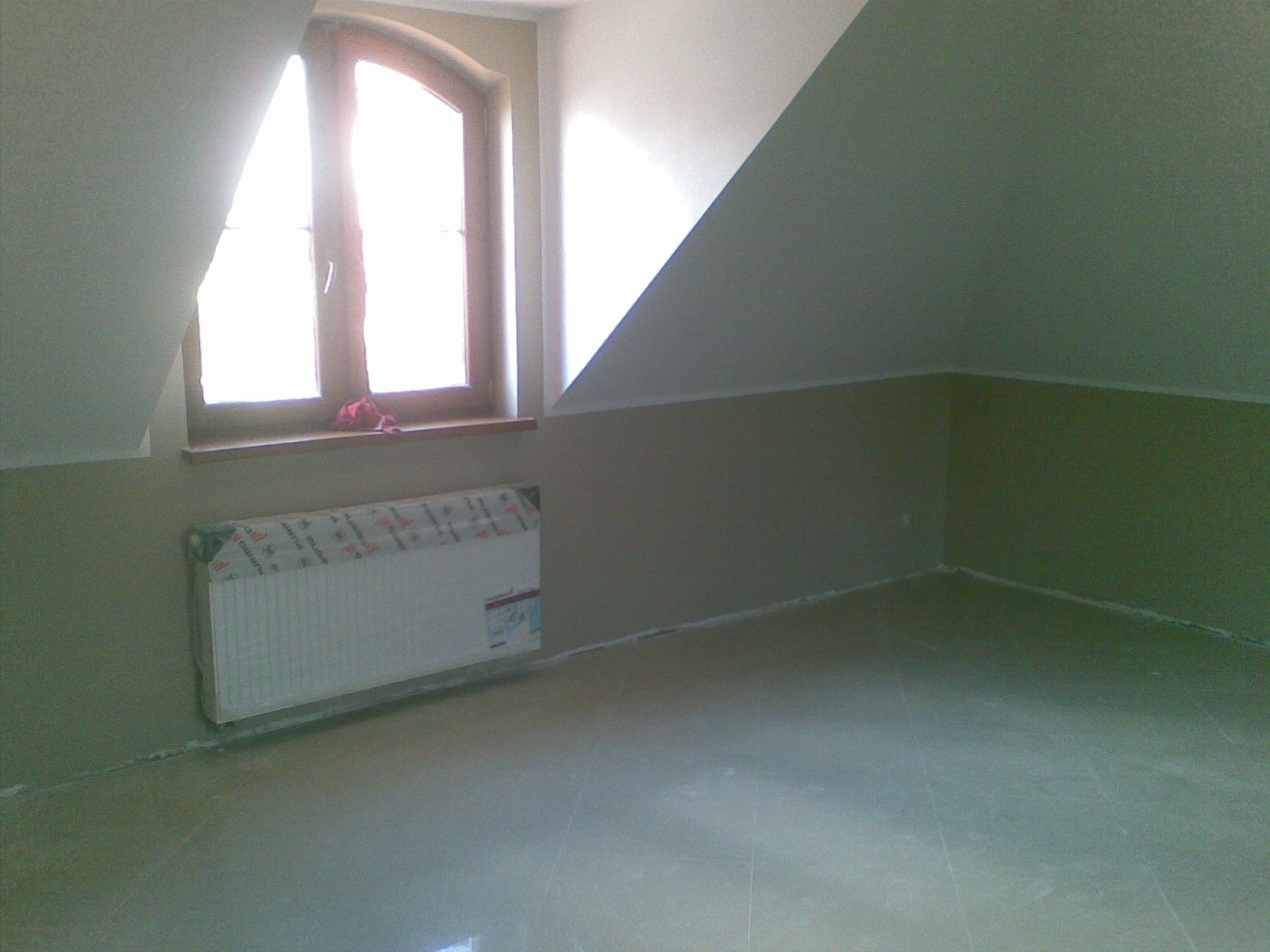 Poddasze po remoncie: świeżo pomalowane ściany w dwóch odcieniach zieleni, okno łukowe z parapetem i zasłonięty grzejnik.