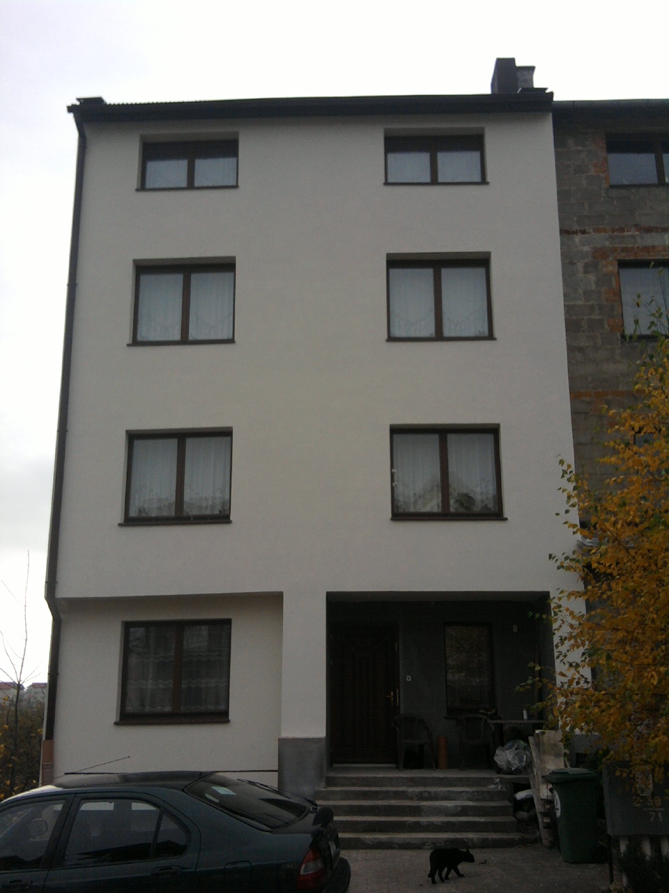 Czteropiętrowy budynek po renowacji elewacji, z widocznym fragmentem sąsiedniego budynku przed renowacją, mały czarny pies przechadza się przed wejściem.