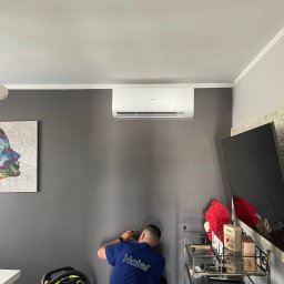 Tb went - Instalator w niebieskiej koszulce z logo Haier wierci otwór w szarej ścianie pod zamontowanym klimatyzatorem Haier; obok stoi odkurzacz Karcher, a w tle widoczny telewizor i dekoracje.