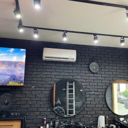 Tb went - Wnętrze salonu fryzjerskiego z czarną ceglaną ścianą, klimatyzatorem Beretta, okrągłymi lustrami i punktowym oświetleniem sufitowym.