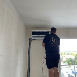 Tb went - Mężczyzna w czarnej koszulce z logo firmy wentylacyjnej stojący na drabinie i montujący klimatyzator ścienny w jasnym pomieszczeniu z widokiem na osiedle domów jednorodzinnych.