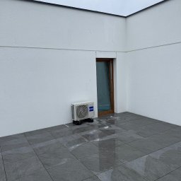 Tb went - Biała jednostka klimatyzacyjna Haier zamontowana na zewnątrz budynku, obok brązowych drzwi na szarym tarasie z płytkami. Widok na pochmurne niebo.