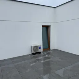 Biała jednostka klimatyzacyjna Haier zamontowana na zewnątrz budynku, obok brązowych drzwi na szarym tarasie z płytkami. Widok na pochmurne niebo.