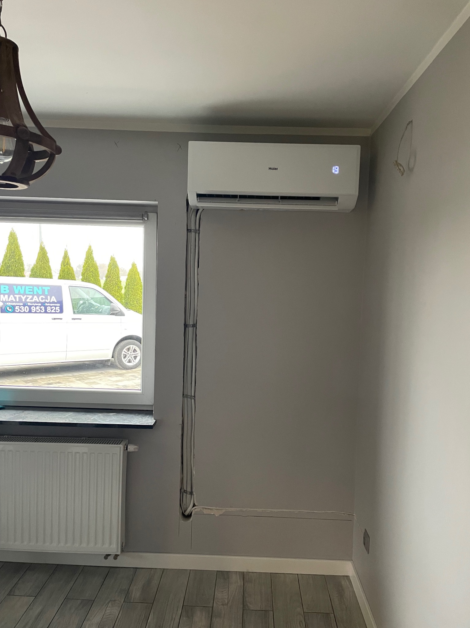Biały klimatyzator Haier zamontowany na szarej ścianie w pomieszczeniu, z widocznymi przewodami w listwie przypodłogowej, okno z widokiem na firmowy samochód z napisem 'B Went Klimatyzacja'.