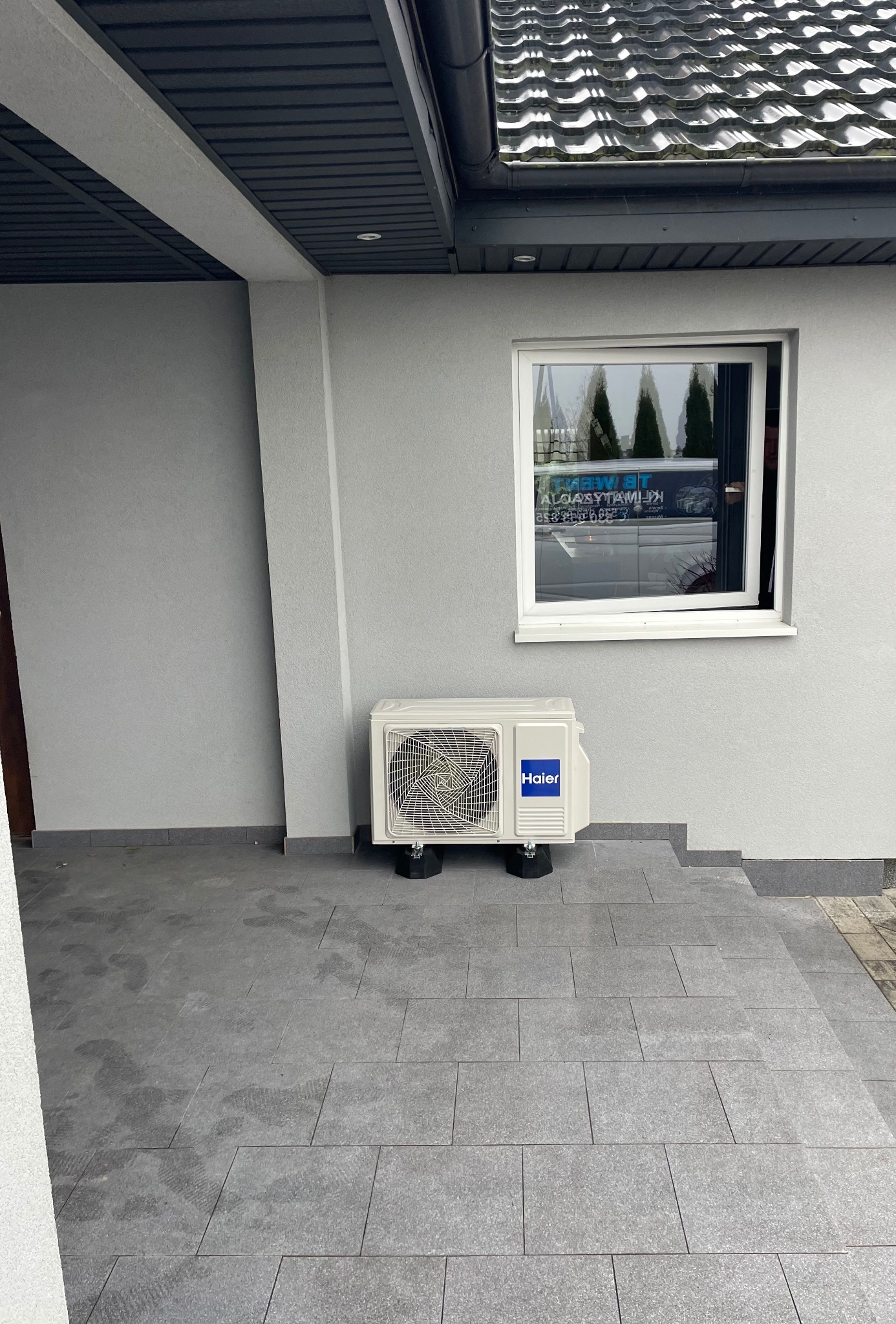 Zewnętrzna jednostka klimatyzacji marki Haier zamontowana na czarnych podkładkach pod oknem w szarej elewacji domu z szarą kostką brukową.