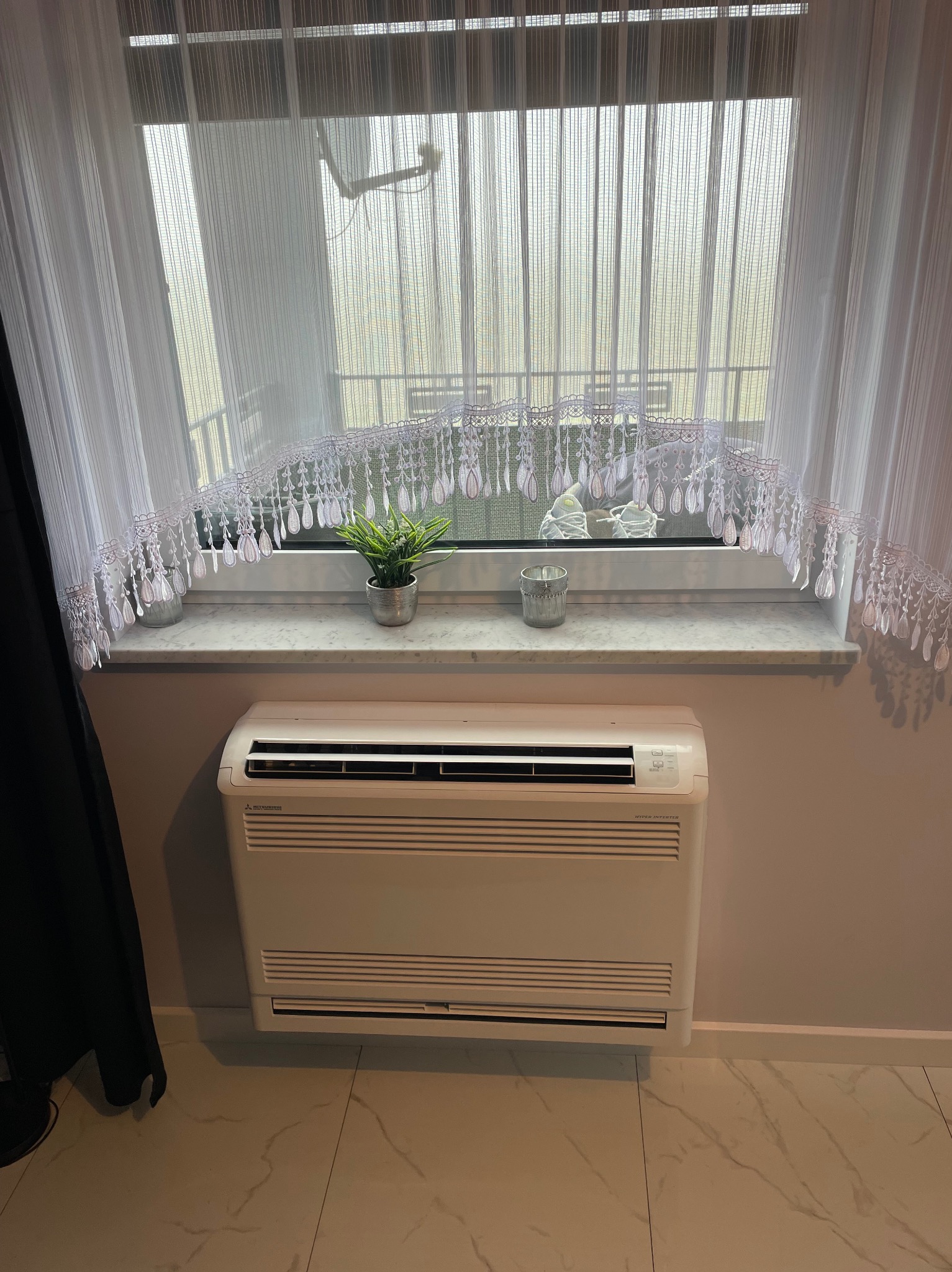 Stojący klimatyzator Mitsubishi Inverter w pomieszczeniu w Złotnikach Kujawskich, z firanką i dekoracjami na parapecie.