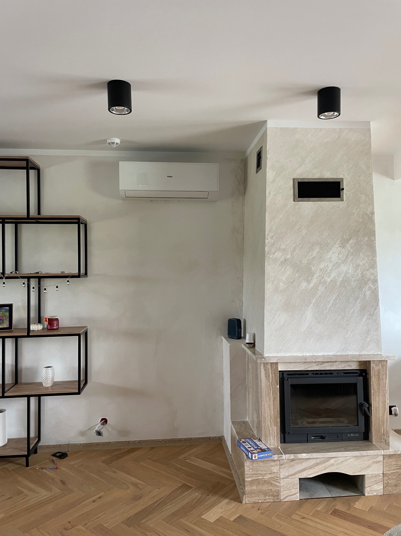 Nowoczesny salon z klimatyzatorem Haier nad kominkiem z marmurową obudową, widoczne również czarne lampy sufitowe i regał z dekoracjami.
