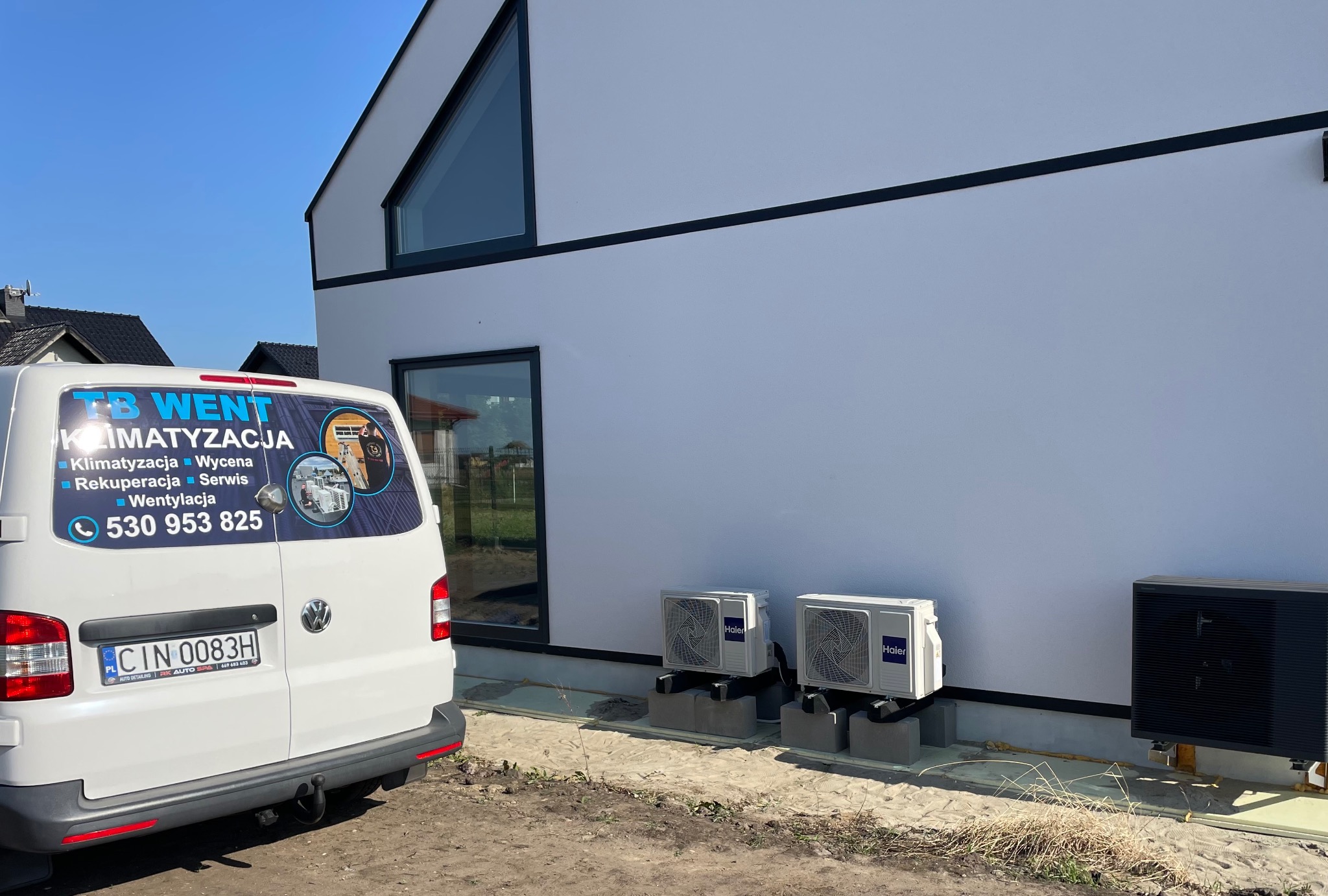 Biały bus firmy TB Went z reklamą usług klimatyzacji i wentylacji zaparkowany obok nowoczesnego, białego domu z zamontowanymi jednostkami zewnętrznymi klimatyzacji Haier na betonowych podstawach.