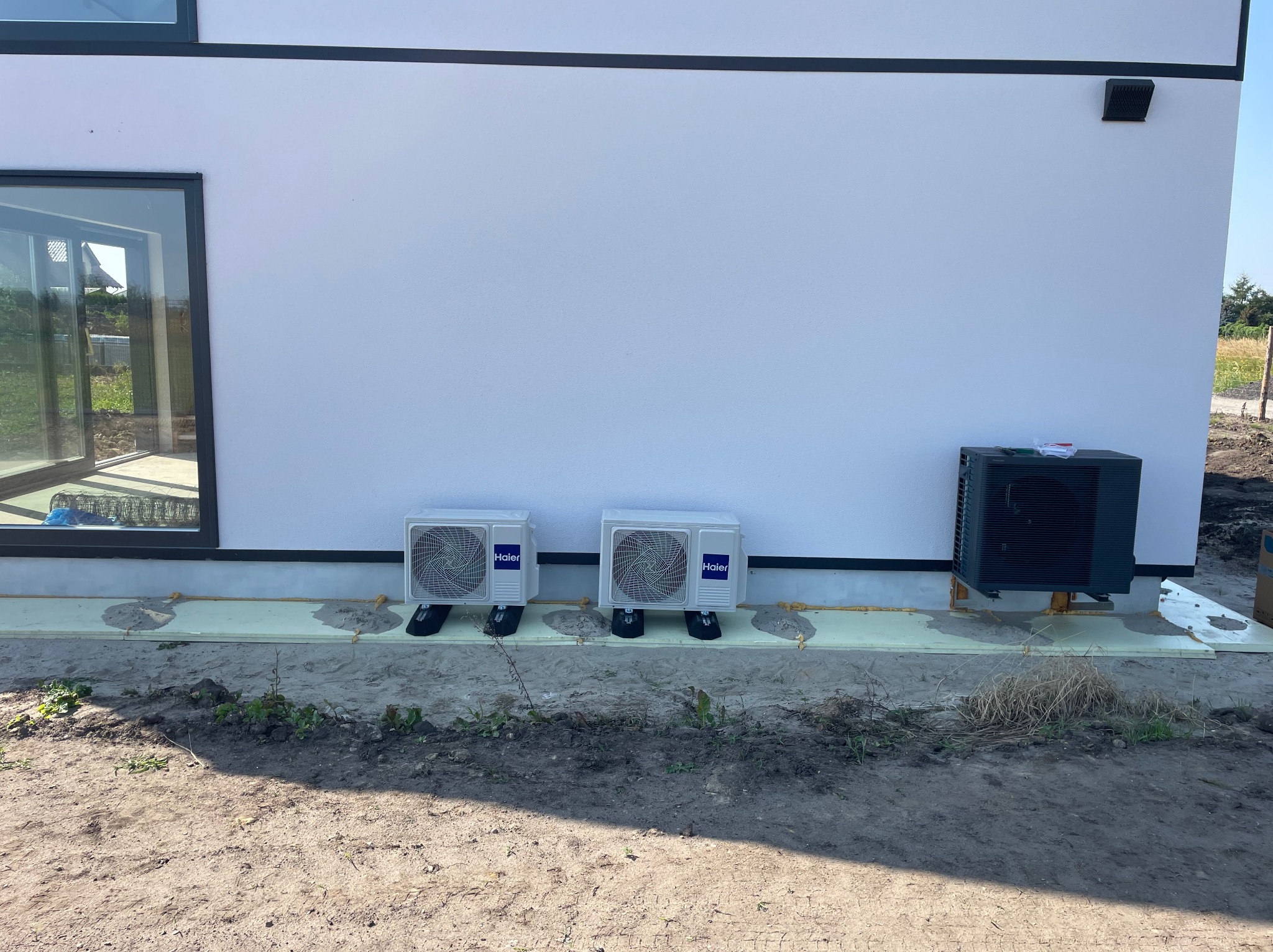 Dwa białe klimatyzatory Haier zamontowane na zewnątrz budynku z jasną elewacją, trzeci, ciemny klimatyzator po prawej, widoczne elementy instalacji i podłoże z piaskiem i resztkami roślinności.