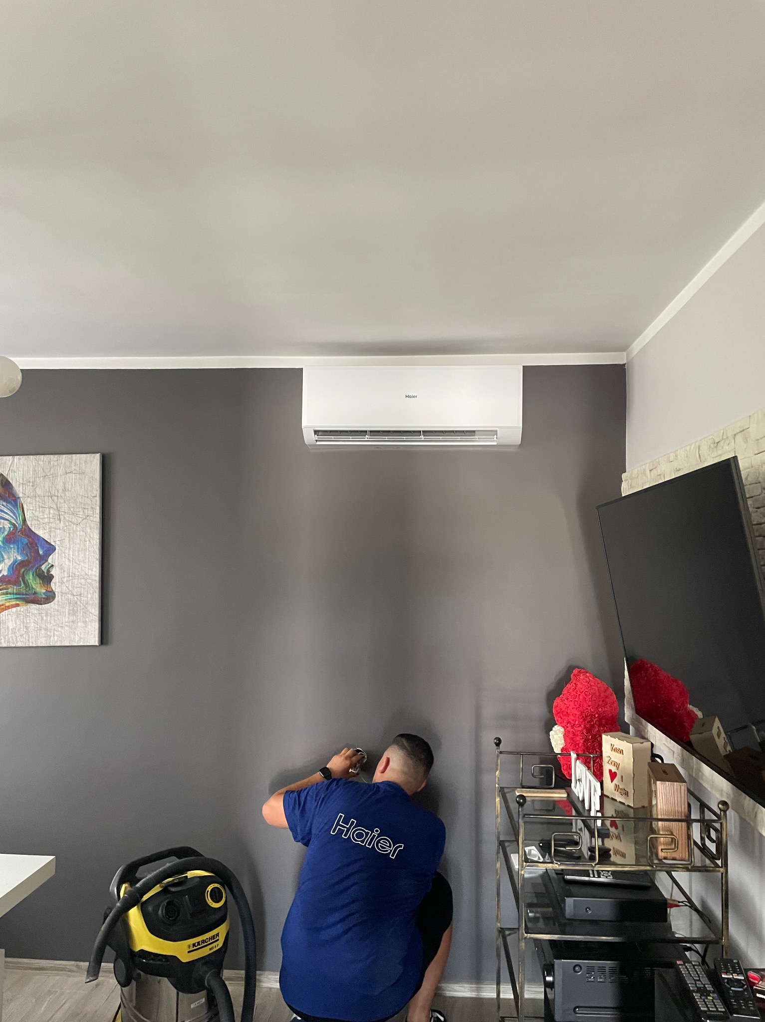 Instalator w niebieskiej koszulce z logo Haier wierci otwór w szarej ścianie pod zamontowanym klimatyzatorem Haier; obok stoi odkurzacz Karcher, a w tle widoczny telewizor i dekoracje.