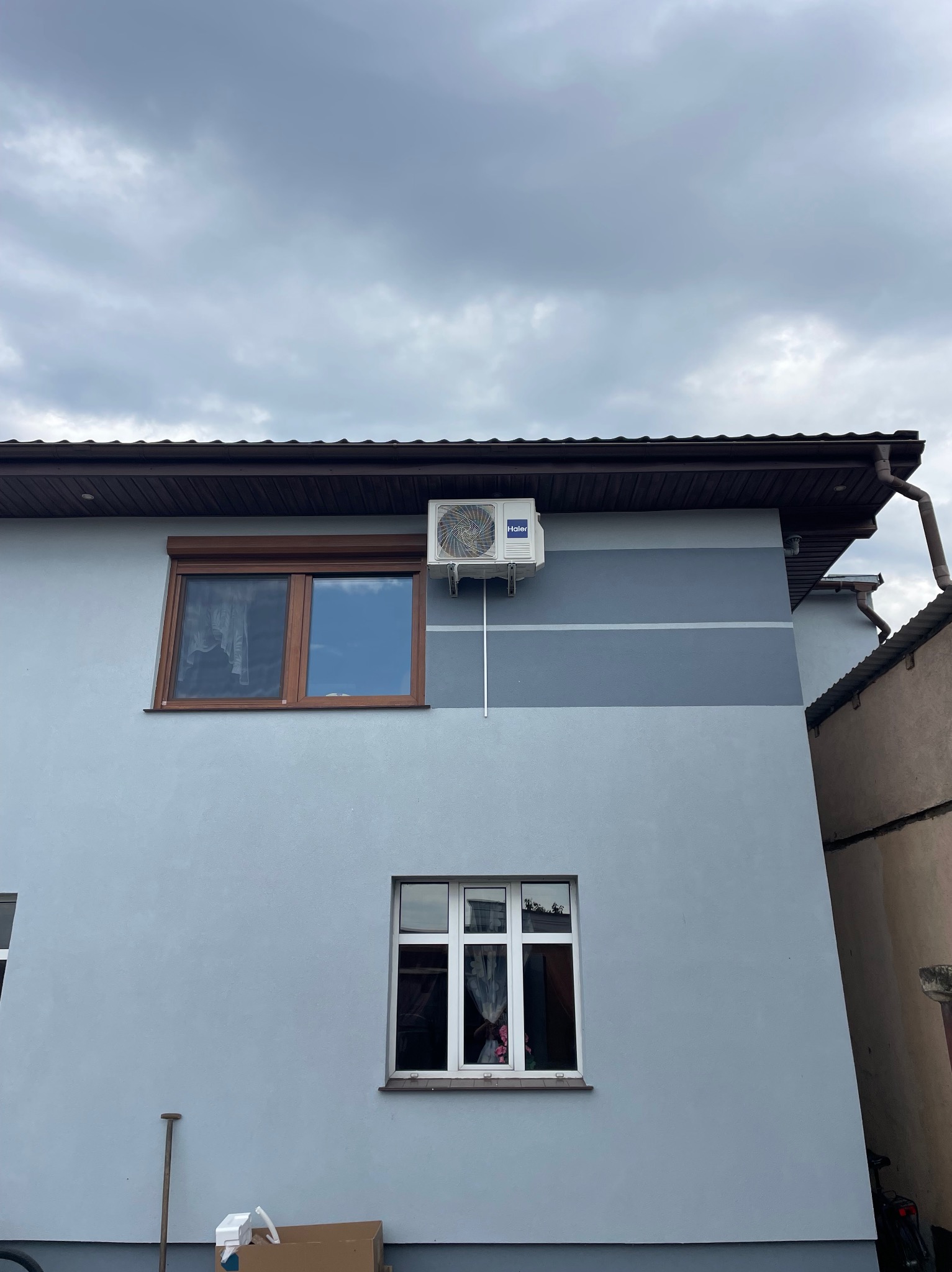 Zewnętrzna jednostka klimatyzacji Haier zamontowana na bocznej ścianie domu z dwoma oknami, widoczne rury odprowadzające, pochmurne niebo w tle.