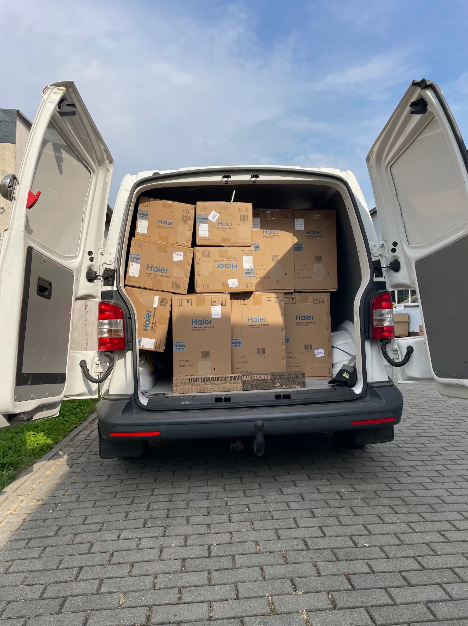 Furgonetka z otwartymi drzwiami załadowana kartonami z klimatyzatorami Haier, widok z tyłu na brukowany podjazd.