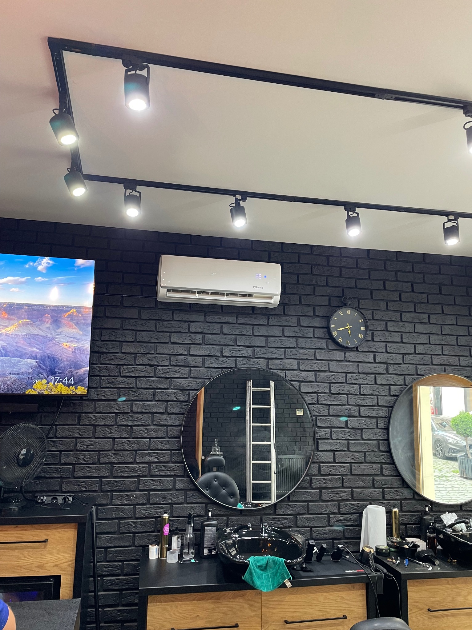 Wnętrze salonu fryzjerskiego z czarną ceglaną ścianą, klimatyzatorem Beretta, okrągłymi lustrami i punktowym oświetleniem sufitowym.