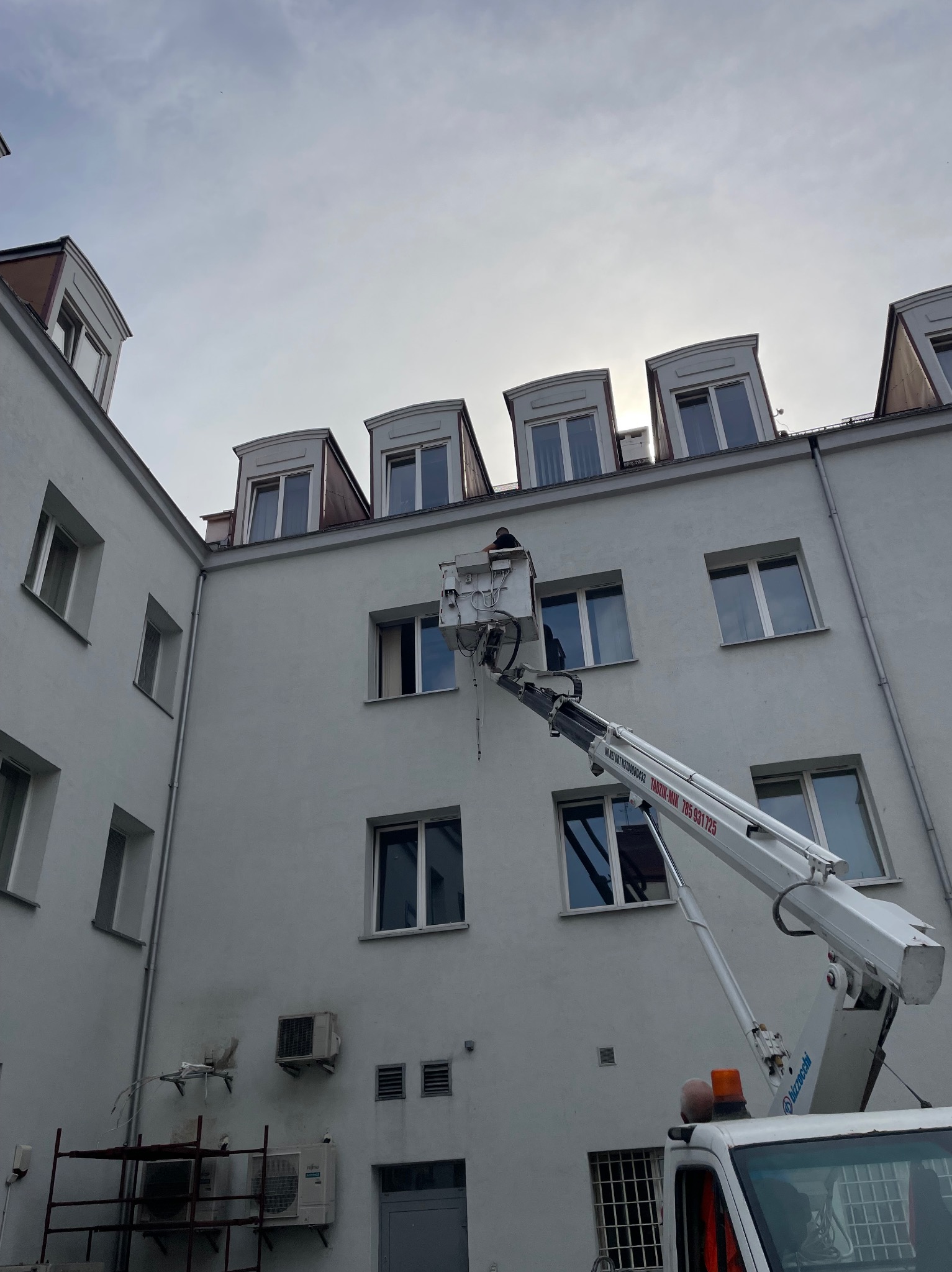 Pracownik na podnośniku koszowym przy elewacji budynku z zamontowanymi klimatyzatorami, widoczne okna dachowe i klimatyzatory naścienne.