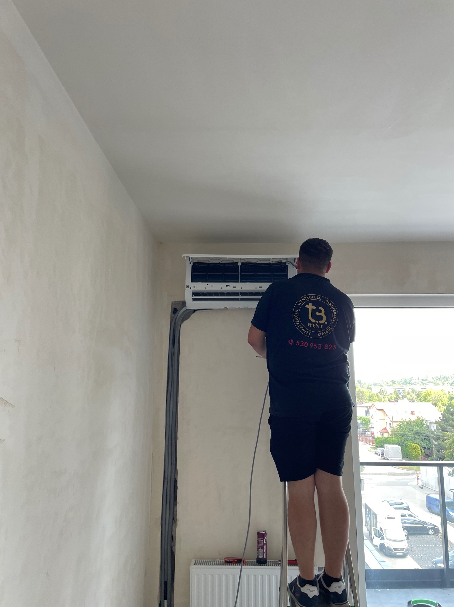 Mężczyzna w czarnej koszulce z logo firmy wentylacyjnej stojący na drabinie i montujący klimatyzator ścienny w jasnym pomieszczeniu z widokiem na osiedle domów jednorodzinnych.