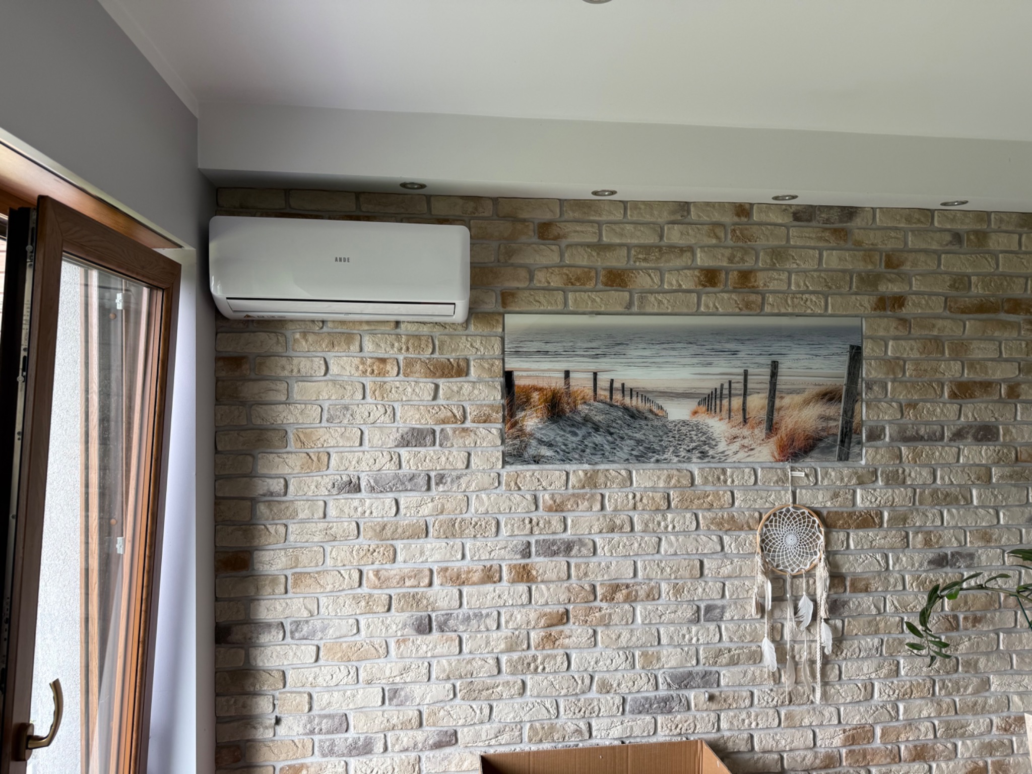 Biały klimatyzator marki ANDE zamontowany na ścianie z cegły dekoracyjnej w salonie. Nad nim sufit z oświetleniem punktowym, obok okno i obraz z motywem plaży.