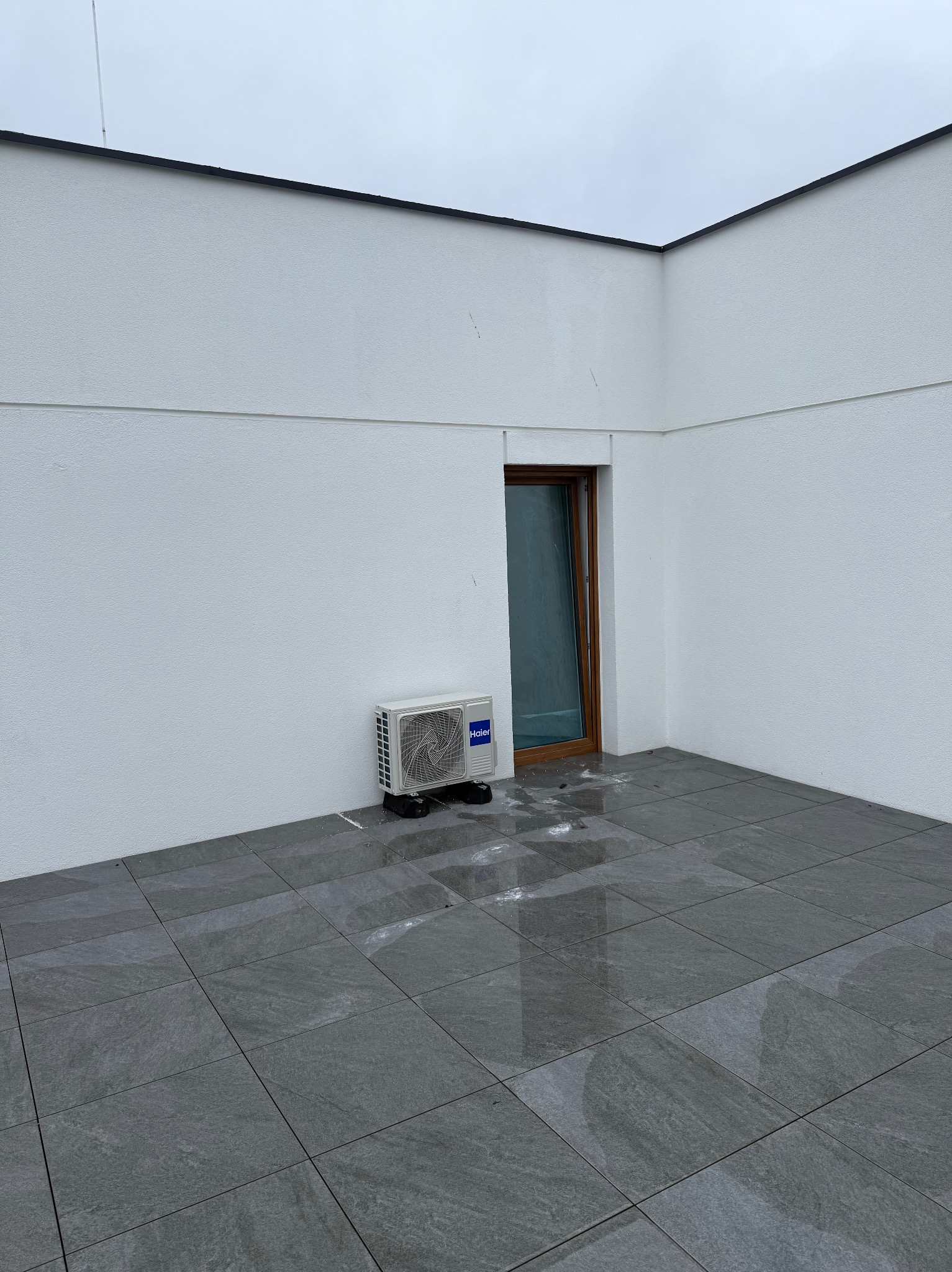 Biała jednostka klimatyzacyjna Haier zamontowana na zewnątrz budynku, obok brązowych drzwi na szarym tarasie z płytkami. Widok na pochmurne niebo.