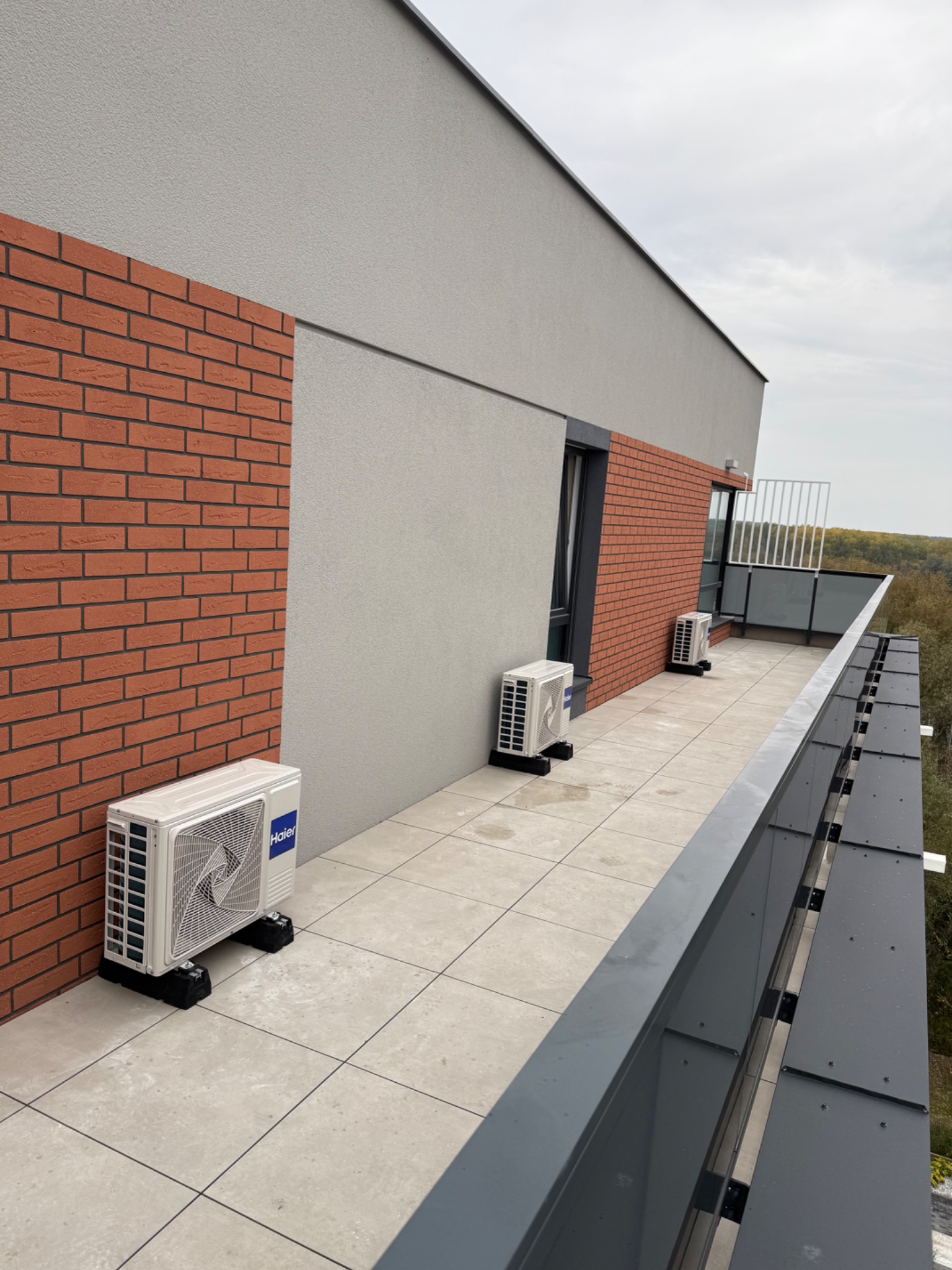 Białe jednostki zewnętrzne klimatyzacji Haier na balkonie nowoczesnego budynku z ceglaną elewacją i szarą ścianą. Widok z góry na zainstalowane urządzenia.